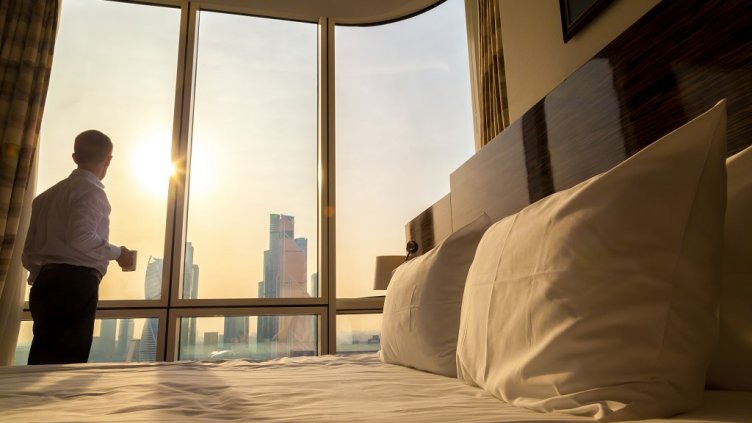 Bed maid-up with white pillows and bed sheets in cozy room. Young businessman with cup of coffee standing at window looking at city scenery on the background. Focus on cushion. Motivation concept