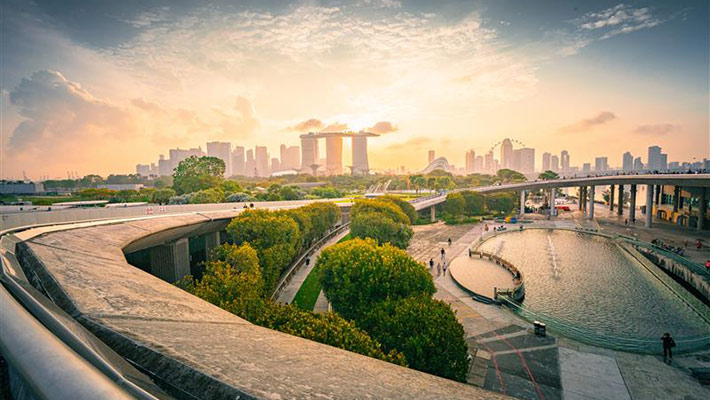 A cityscape view of Singapore at sunset featuring Marina Bay Sands, lush greenery, footpaths, and a curved water feature in the foreground.