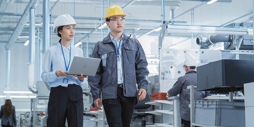 Two Young Asian Heavy Industry Engineers in Hard Hats Walking with Laptop Computer and Discussing Work Process in a Factory